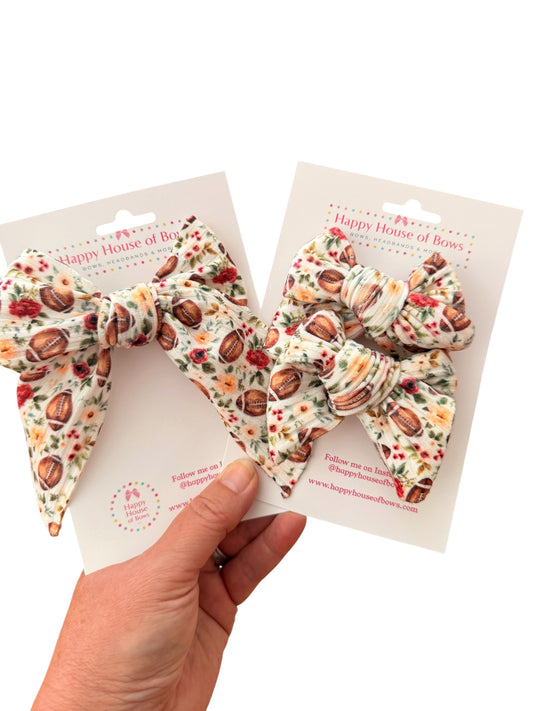 Two fabric hair bows with a floral and football print, attached to white display cards and held by a hand against a white background.