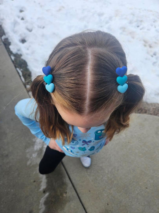 Child with pigtails and heart-shaped hair clips standing on a snowy sidewalk.