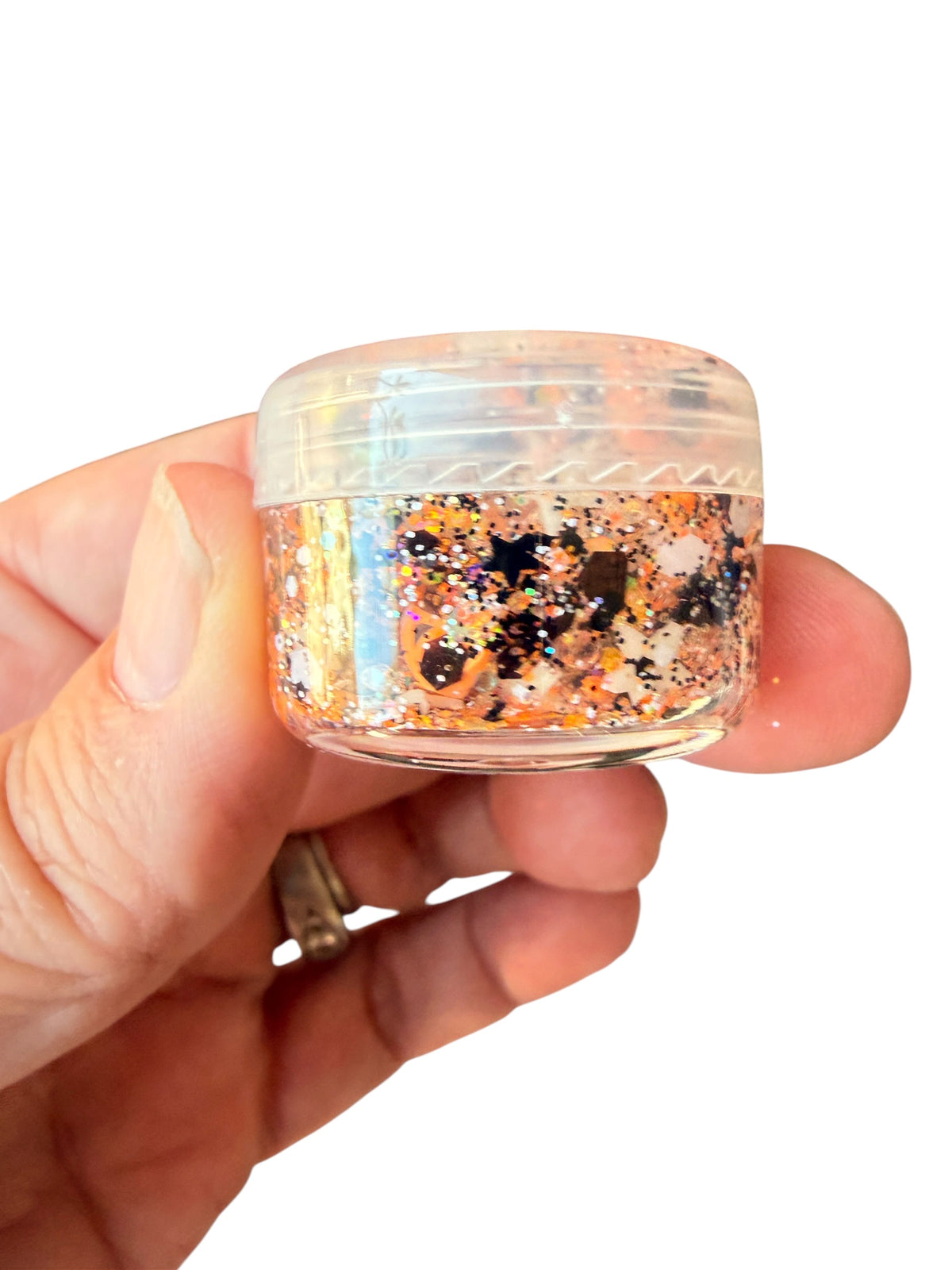 A hand holding a small clear plastic jar filled with iridescent hair gel glitter in orange, gold, black, and silver tones against a plain white background.