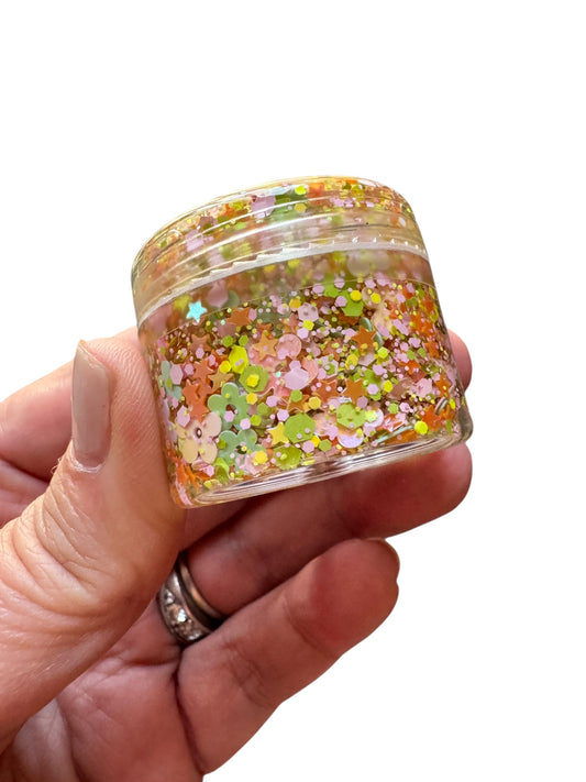 Hand holding a small jar of colorful glitter hair and body gel against a white background