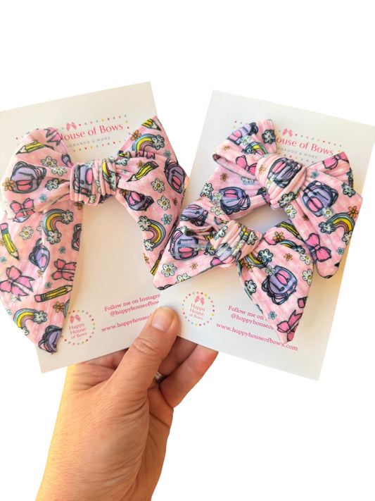 Two pink fabric hair bows with a colorful cartoon pattern are displayed on white cards held by a person's hand against a white background.