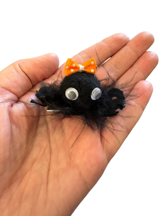 A black fuzzy hair clip shaped like a spider with white eyes and an orange bow on top in a hand
