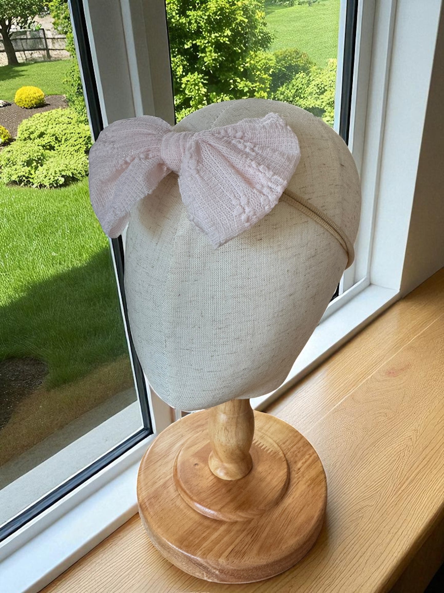 Mannequin head with a pink knitted bow on a wooden stand by a window with a view of greenery.