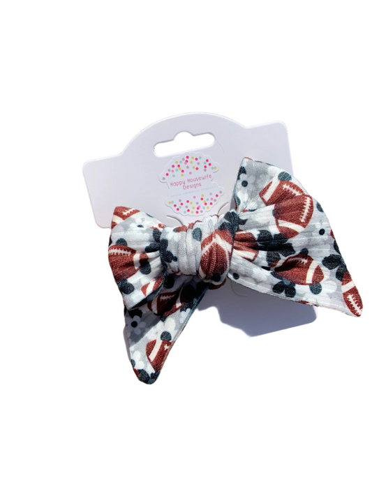A hand holding a hair bow with a floral and football pattern on it, displayed against a grassy background.