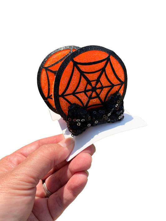 A pair of orange and black sequin mouse ear clips with a spiderweb pattern and black sequin bows, displayed on a card