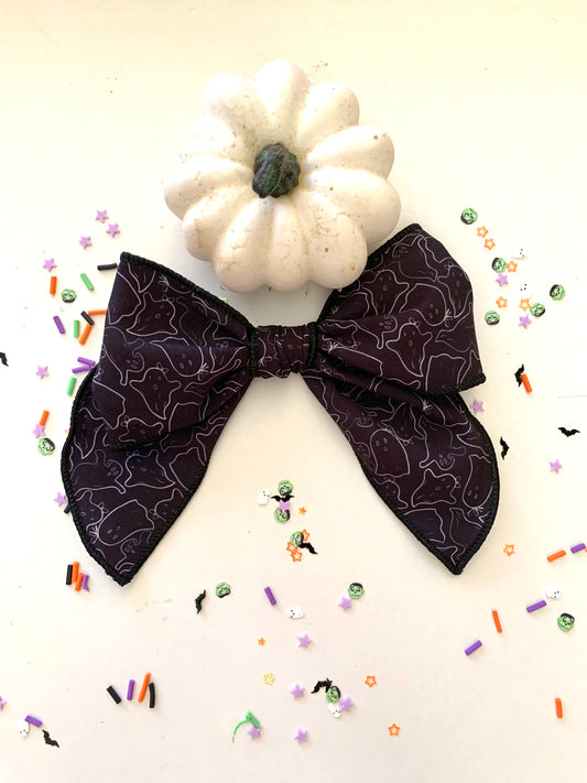 A black hair bow with a pattern resembling a ghost outline, presented on a white surface with a small white pumpkin and decorative sprinkles around it.