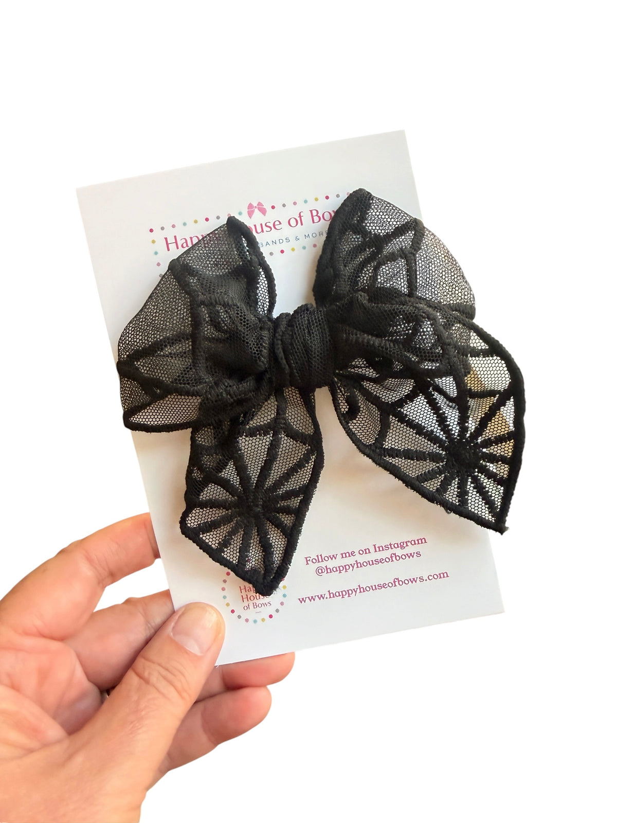 A black sheer lace bow with a spiderweb pattern, held on a white display card by a hand against a white background.