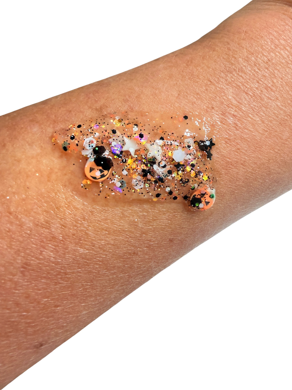 A close-up of a person's forearm with a dollop of clear hair and body gel containing multicolor glitter and small decorative pieces, shown against a plain white background.