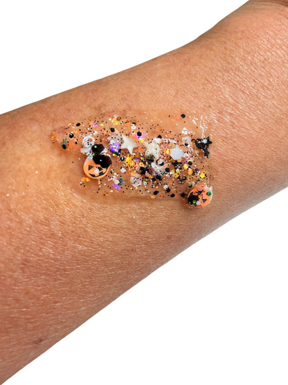 A close-up of a person's forearm with a dollop of clear hair and body gel containing multicolor glitter and small decorative pieces, shown against a plain white background.