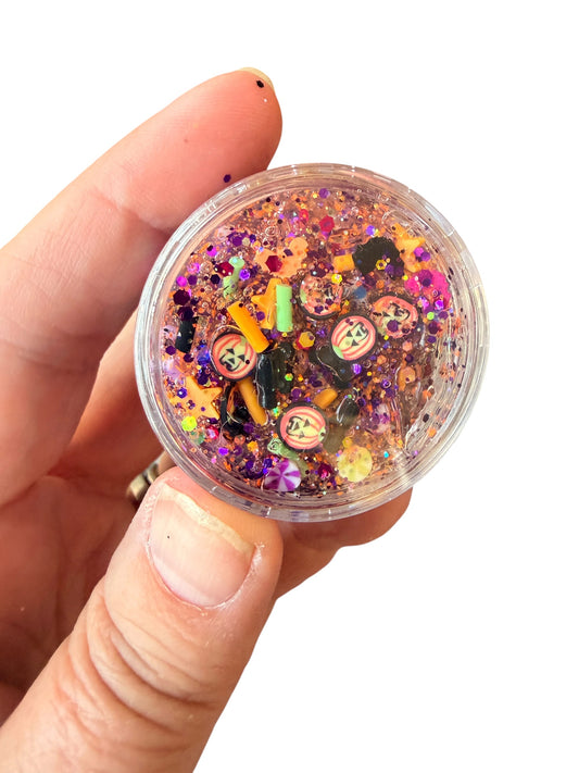 A hand holding a small clear round container filled with colorful glitte rhair gel, sequins, and small decorative charms against a plain white background.