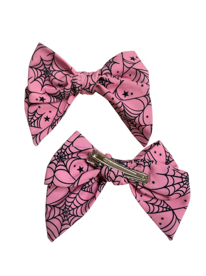 Two pink hair bows with a black spiderweb and star pattern, one shown flat and the other attached to a silver metal hair clip, on a plain white background.