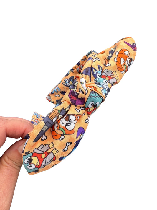 A headband with a pattern of blue dogs and Halloween-themed elements on a yellow background, attached to an alligator clip.