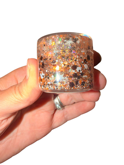 A hand holding a small clear jar filled with shimmering orange glitter hair gel against a plain white background.