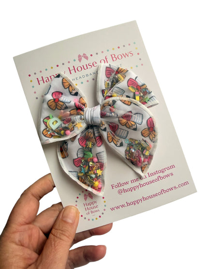 A large fabric hair bow with a colorful butterfly and sewing machine pattern on a white background, held by a hand against a white backdrop. The bow is attached to a branded display card for 'Happy House of Bows'.