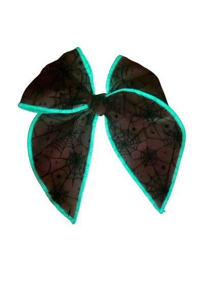 A large hair bow featuring a black spiderweb pattern on a sheer mesh fabric, finished with a glow in the dark trim.