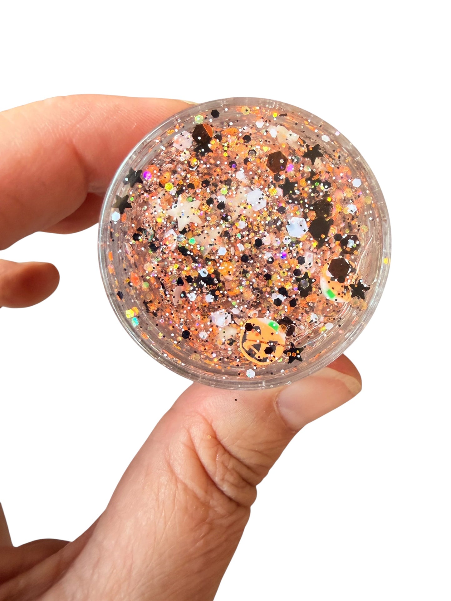 A hand holding a small clear round container filled with multicolor hair gel glitter, including gold, orange, and black flakes, against a plain white background.