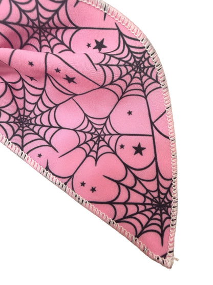 A close-up of a pink hair bow with a black spiderweb and star pattern, finished with a light pink fringed edge, set against a plain white background.