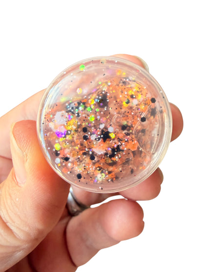 A hand holding a clear round container filled with orange glitter, black flecks, and multicolored holographic hair gel glitter against a white background.