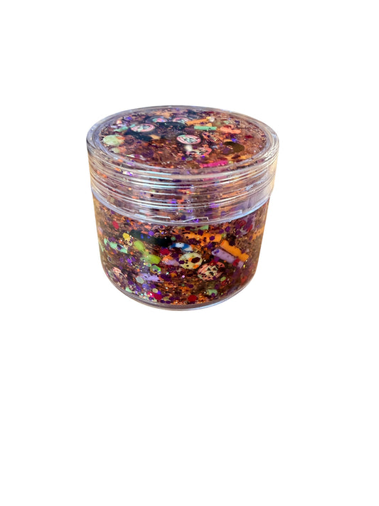 A clear plastic jar filled with Halloween  glitter hair gel and decorated with colorful confetti, sequins, and small skull embellishments, shown on a plain white background.