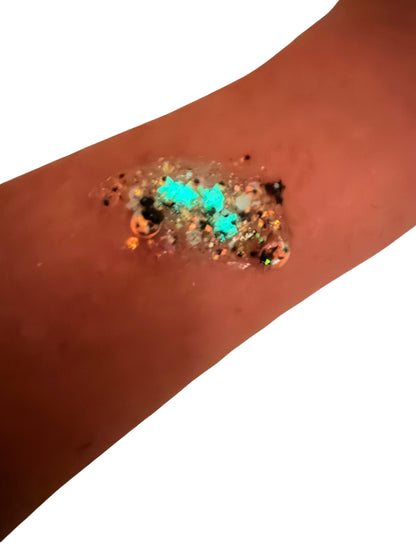 A close-up of a forearm with a patch of hair or body gel featuring bright turquoise, black, orange, and glittery glow in the dark details on a plain white background.