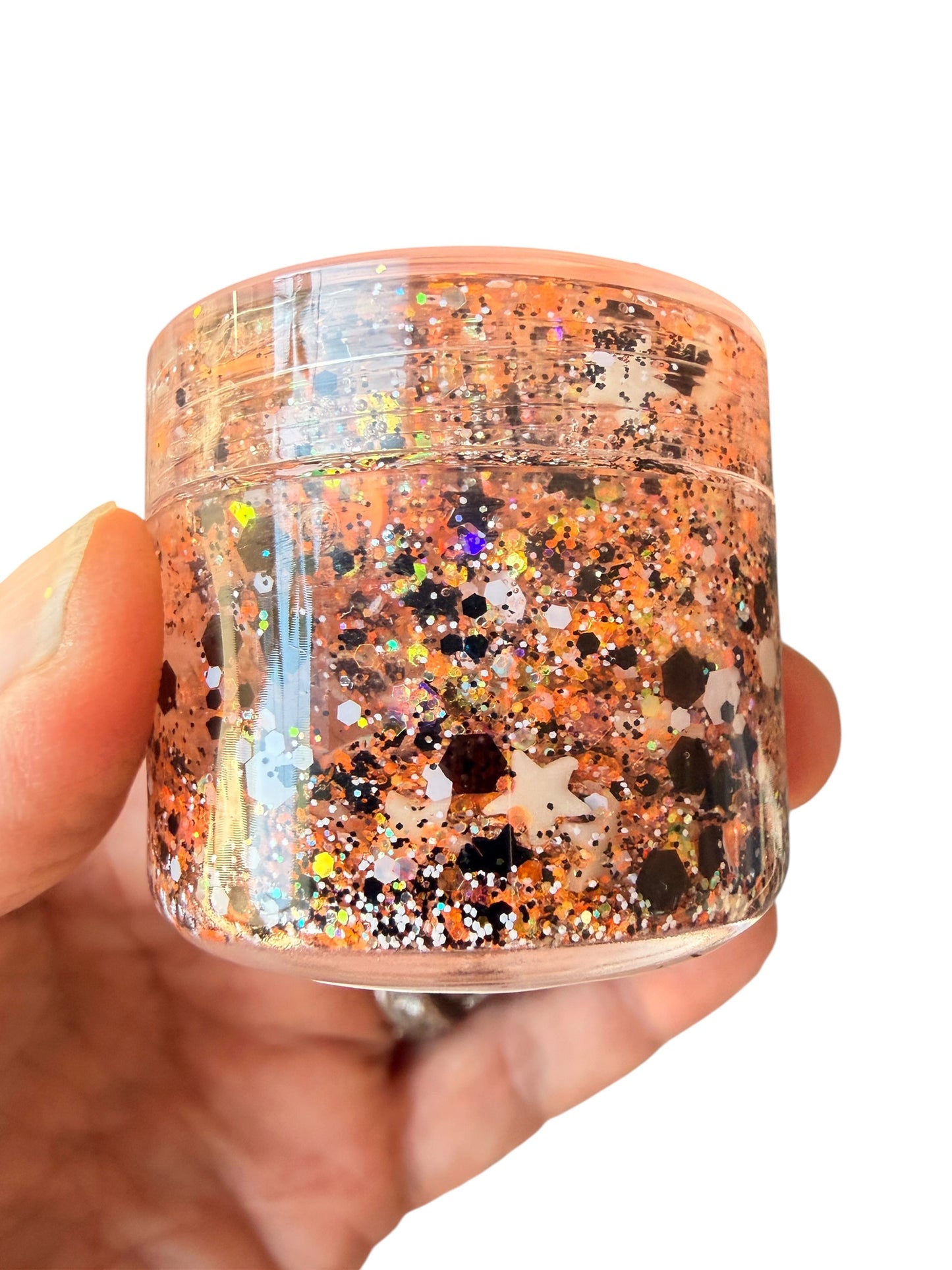 A hand holding a small clear plastic jar of hair gel filled with shimmering orange, gold, and black glitter against a plain white background.