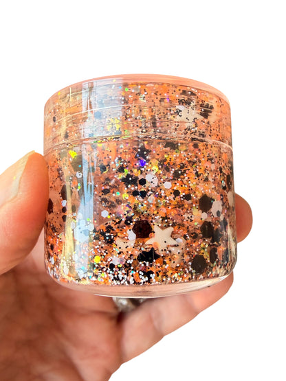 A hand holding a small clear plastic jar of hair gel filled with shimmering orange, gold, and black glitter against a plain white background.