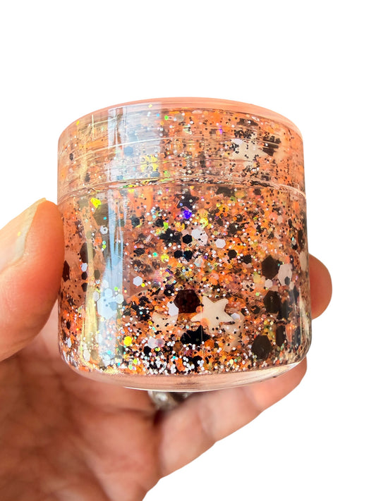 A hand holding a small clear plastic jar of hair gel filled with shimmering orange, gold, and black glitter against a plain white background.
