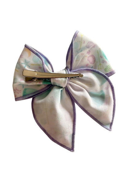 A fabric hair bow clip featuring a pastel multicolor mermaid-themed print with purple stitched edges and a gold-tone metal clip, shown on a plain white background.