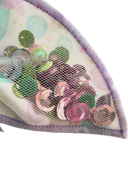 Close-up of a mesh garment trim with pastel polka dots and shiny multicolored sequins, finished with a lavender stitched edge on a white background.