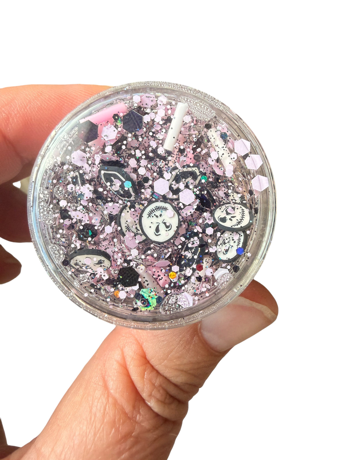 A hand holding a small clear round container of halloween hair gel glitter filled with pink, black, and white glitter, flakes, and decorative charms against a plain white background.