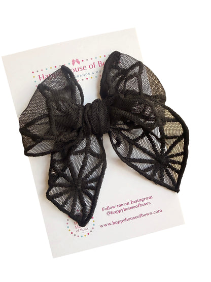 A large black lace bow with a spiderweb pattern, displayed on a white branded card from Happy House of Bows.