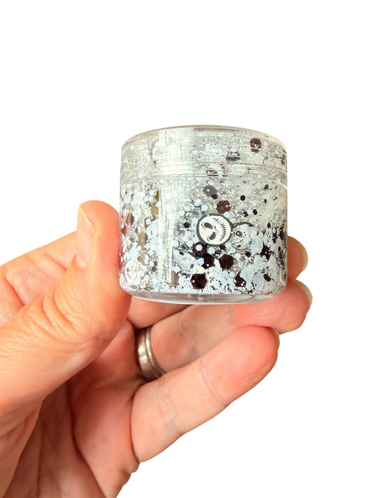 A hand holding a small clear jar filled with black and white hair gel glitter and black speckles against a plain white background.
