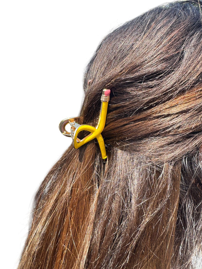 Hair clip shaped like a yellow pencil with pink eraser on brown hair against a white background