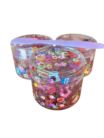 Three clear plastic jars filled with pink glitter and decorated with colorful flower-shaped embellishments, with a purple plastic spoon resting across the top, set against a plain white background.