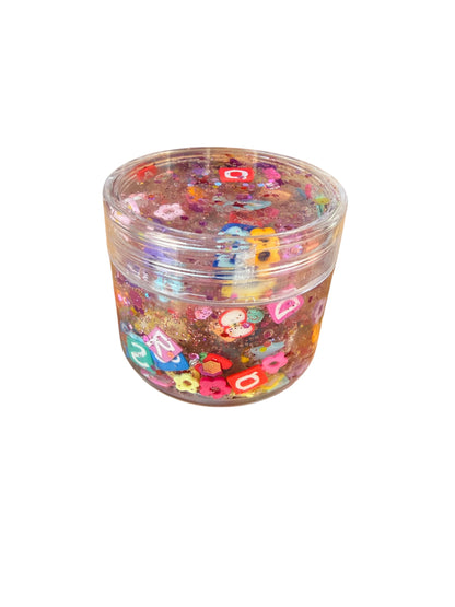 A clear plastic jar filled with colorful glittery slime and decorated with small pastel letter and flower charms, shown on a plain white background.