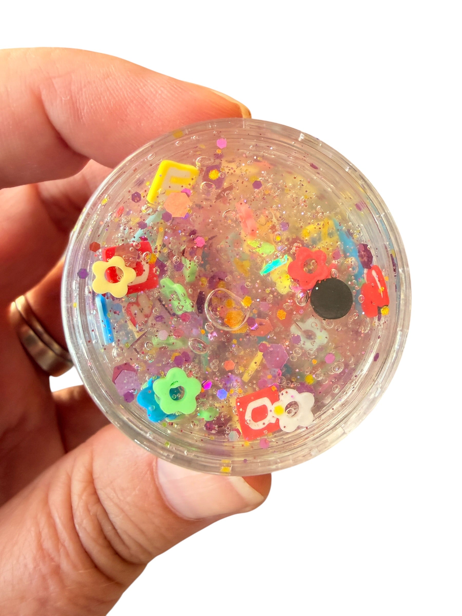 A hand holding a round clear plastic container filled with glittery gold slime and colorful decorative charms, shown against a plain white background.