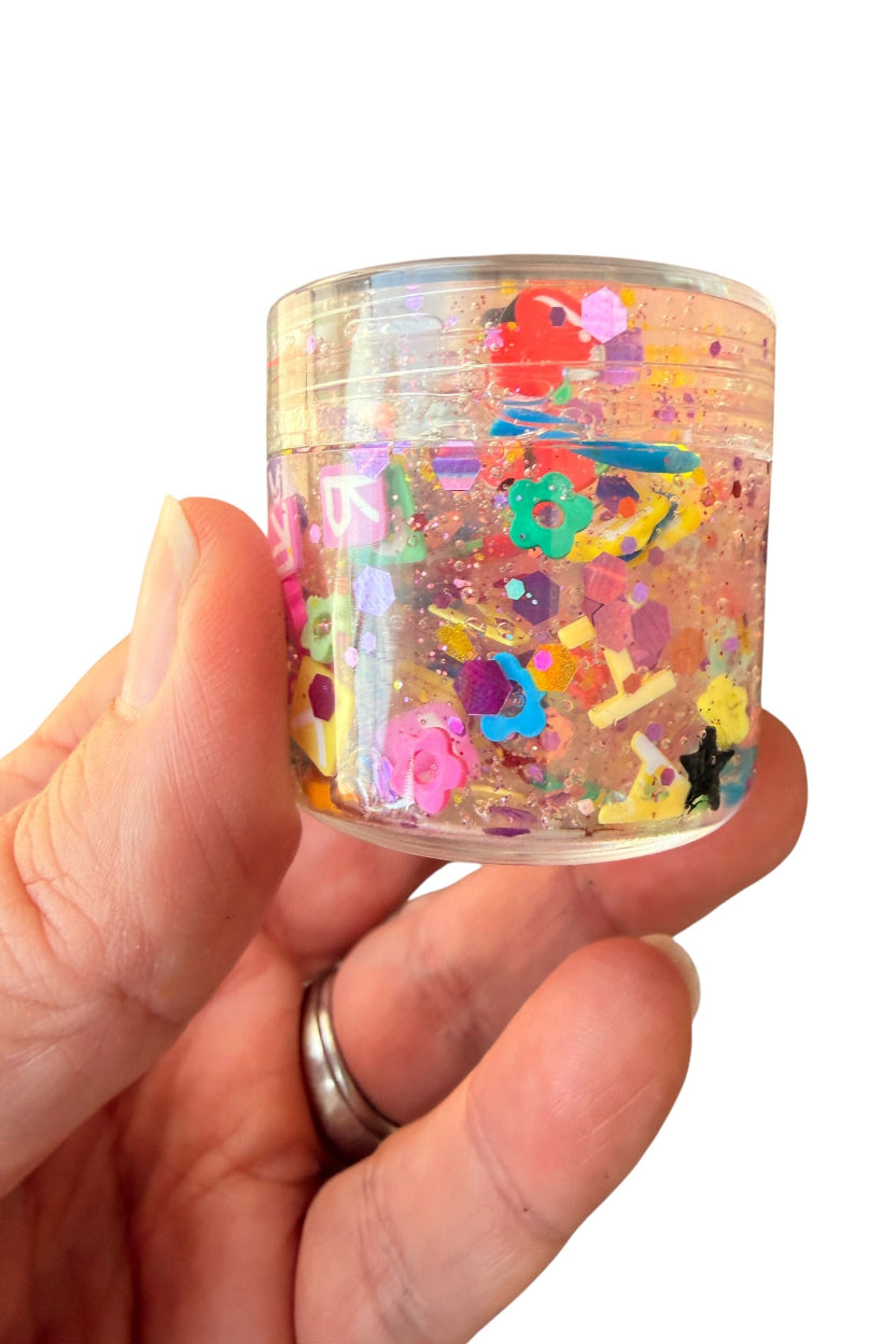 A hand holding a small clear plastic jar filled with colorful glitter, confetti, and small decorative pieces, shown against a plain white background.