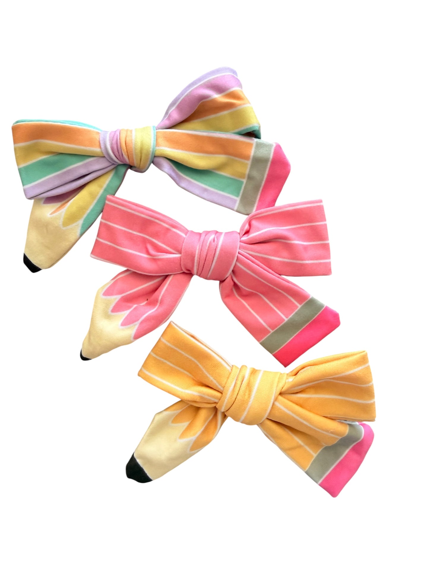 Three colorful bow hair clips designed to look like pencils, arranged diagonally on a solid white background. The bows feature pastel stripes in pink, yellow, orange, green, and purple, with black tip details.