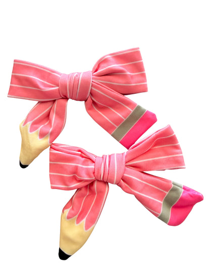 Two pink striped fabric hair bows designed to look like pencils, with yellow erasers and gray graphite accents, shown on a plain white background.