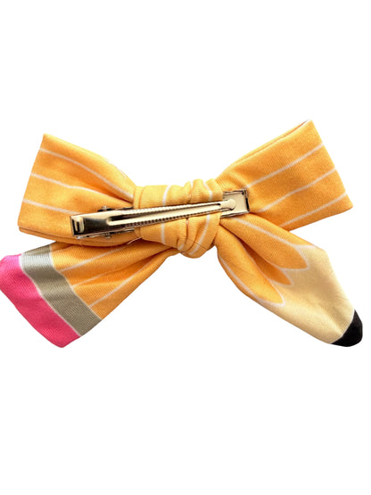 A novelty hair bow designed to look like a pencil, featuring a yellow and cream striped body, a pink eraser tip, and a silver metal claw clip.