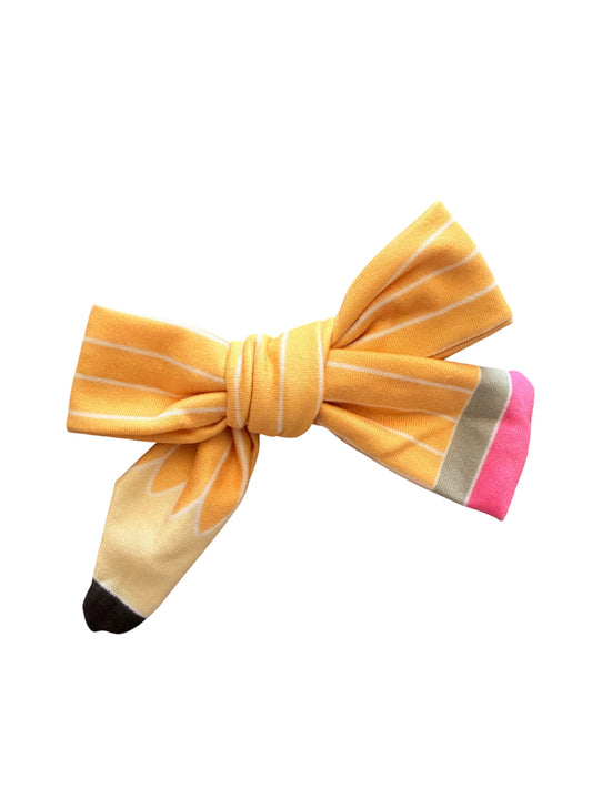 A novelty hair bow designed to look like a pencil, featuring a yellow and cream striped body, a black tip, and a pink eraser end, set against a plain white background.
