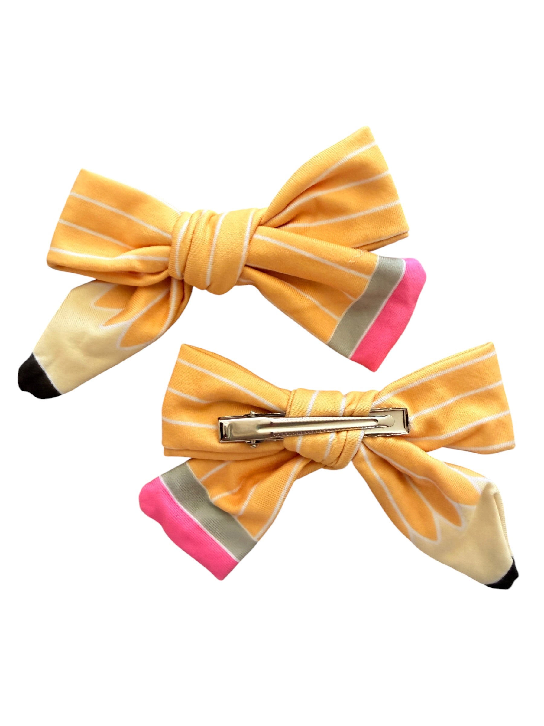Two yellow bow hair clips designed like pencils, featuring a cream-colored wood-grain texture, pink and gray accents, and a silver metal clip, shown on a plain white background.