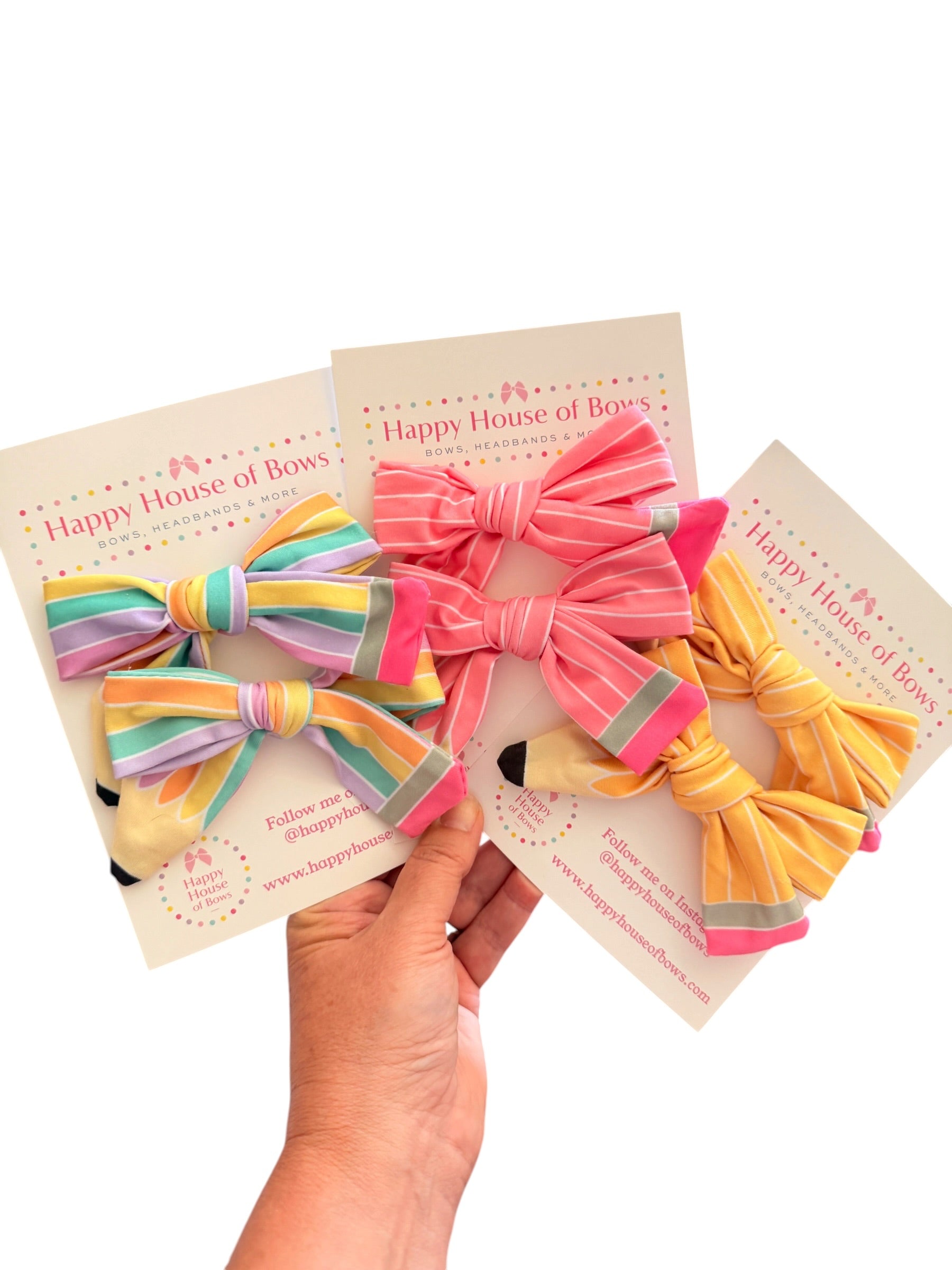 Three striped fabric hair bows on display cards, held by a hand against a white background. The bows are in pastel rainbow, pink, and yellow-orange stripes, with the brand name 'Happy House of Bow' visible on the cards.