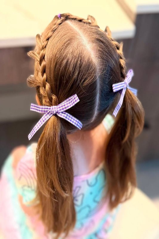 A close-up back view of a child with brown hair styled in two braided pigtails, each tied with a purple and white gingham ribbon bow. The child is wearing a pastel tie-dye shirt in an indoor setting.