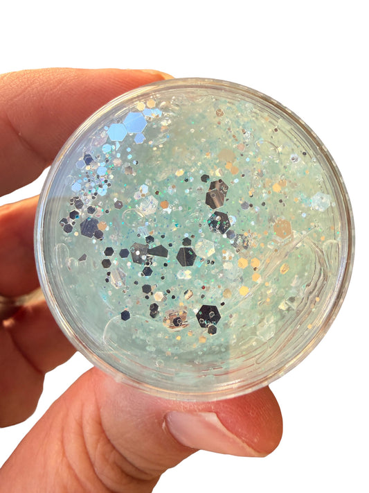A hand holding a small round clear container filled with iridescent glitter  hair gel silver tones with scattered reflective flakes.