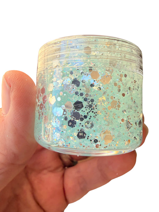 A hand holding a small clear plastic jar filled with iridescent silver hair gel  and scattered metallic hexagonal glitter, against a plain white background.