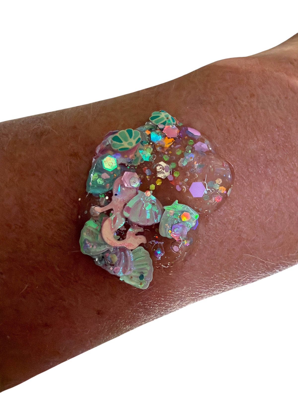 A close-up of a person's forearm with a decorative hair and body gel glitter. The design features iridescent seashell and coral-like motifs with holographic glitter on a clear gel base, shown against a plain white background.